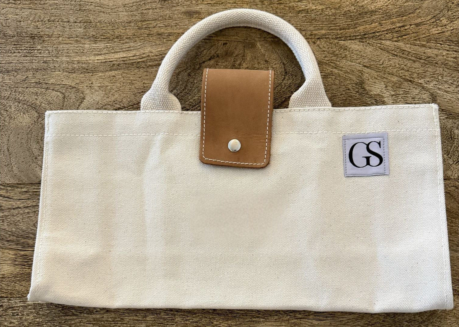 Beige handbag with brown leather patch and 'GS' logo on a wooden surface