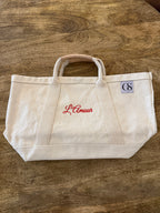 An off-white canvas tote bag with red embroidered 'L'Amour' script on the front and a small blue 'GS' patch on the side, resting on a wooden tabletop.