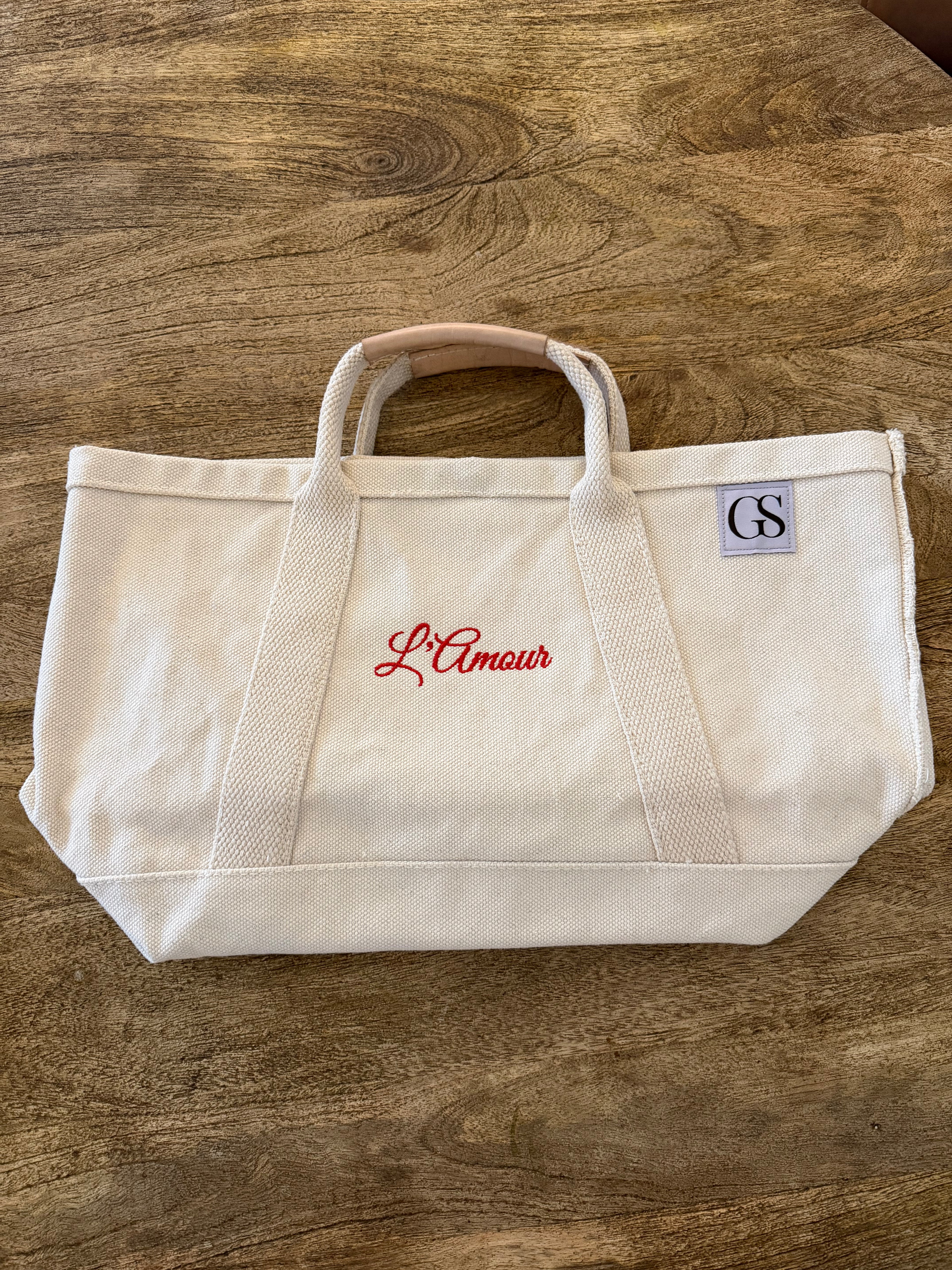 An off-white canvas tote bag with red embroidered 'L'Amour' script on the front and a small blue 'GS' patch on the side, resting on a wooden tabletop.