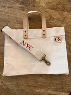 A cream canvas tote bag with light tan leather handles and a matching canvas shoulder strap featuring red embroidered 'NYC' lettering, laid flat on a brown wooden surface. A small square leather patch on the right side of the bag displays 'GS'.