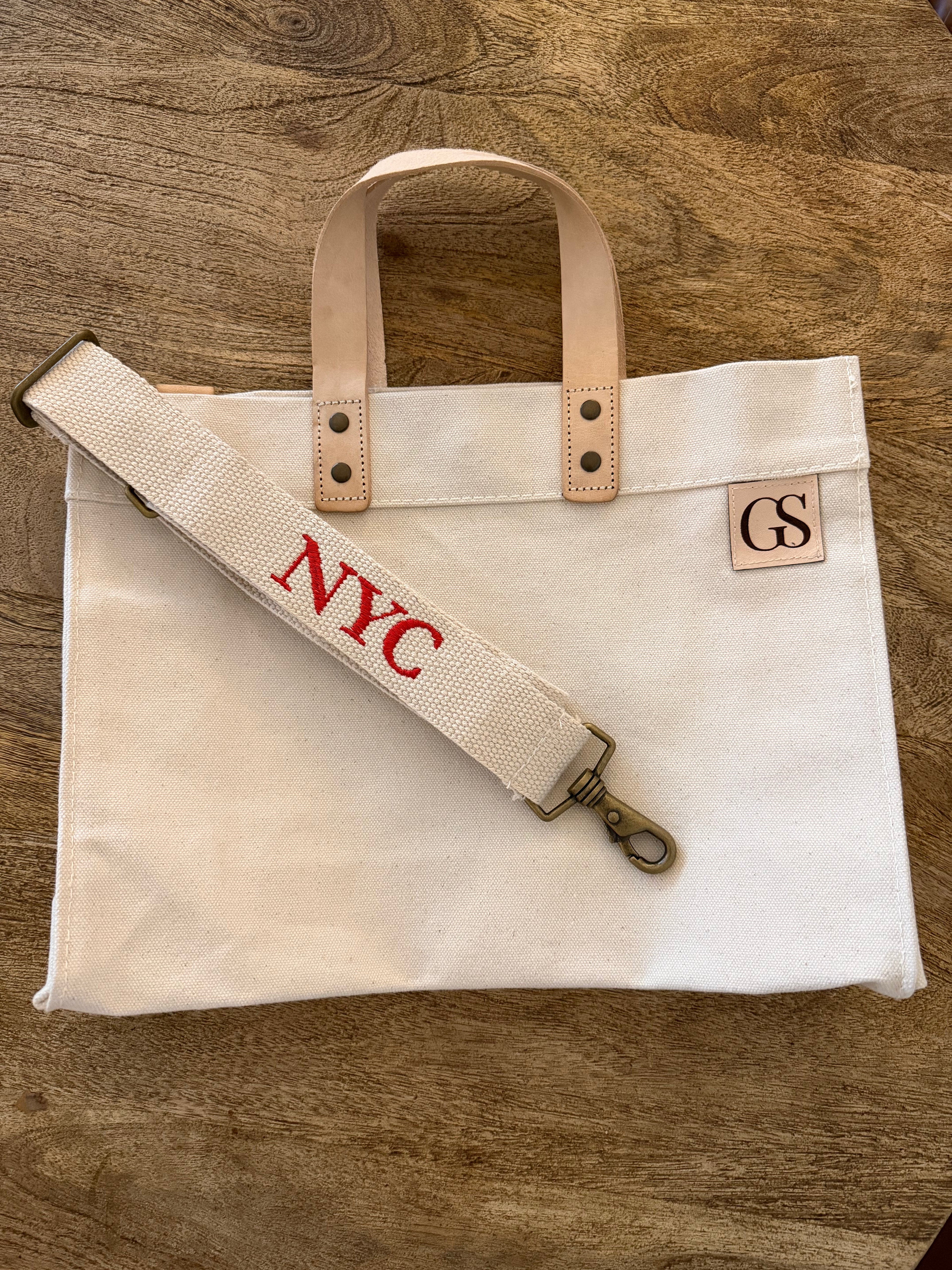 A cream canvas tote bag with light tan leather handles and a matching canvas shoulder strap featuring red embroidered 'NYC' lettering, laid flat on a brown wooden surface. A small square leather patch on the right side of the bag displays 'GS'.
