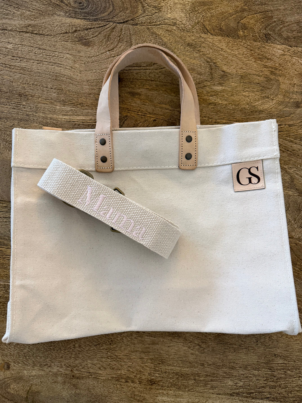 A cream canvas tote bag with light tan leather handles and a matching canvas shoulder strap, displayed on a rustic wooden background. The canvas features a small tan square patch with the monogram 'GS' and the strap is embroidered with the name 'Mara'.