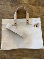 A cream canvas tote bag with light tan leather handles and a matching canvas shoulder strap, displayed on a rustic wooden background. The canvas features a small tan square patch with the monogram 'GS' and the strap is embroidered with the name 'Mara'.