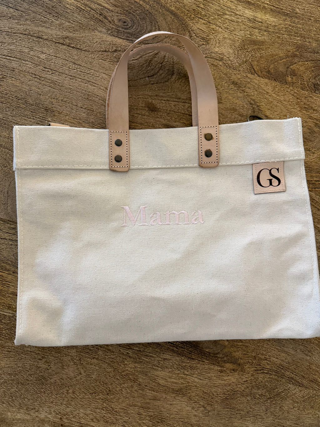 An off-white canvas tote bag with light tan leather handles and a small square leather patch with the initials 'GS'. The bag has 'Mama' embroidered in light pink across the front, and is photographed flat on a brown wood-grain surface.
