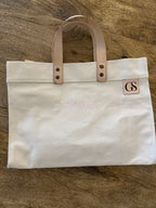 An off-white canvas tote bag with light tan leather handles and a small square leather patch with the initials 'GS'. The bag has 'Mama' embroidered in light pink across the front, and is photographed flat on a brown wood-grain surface.