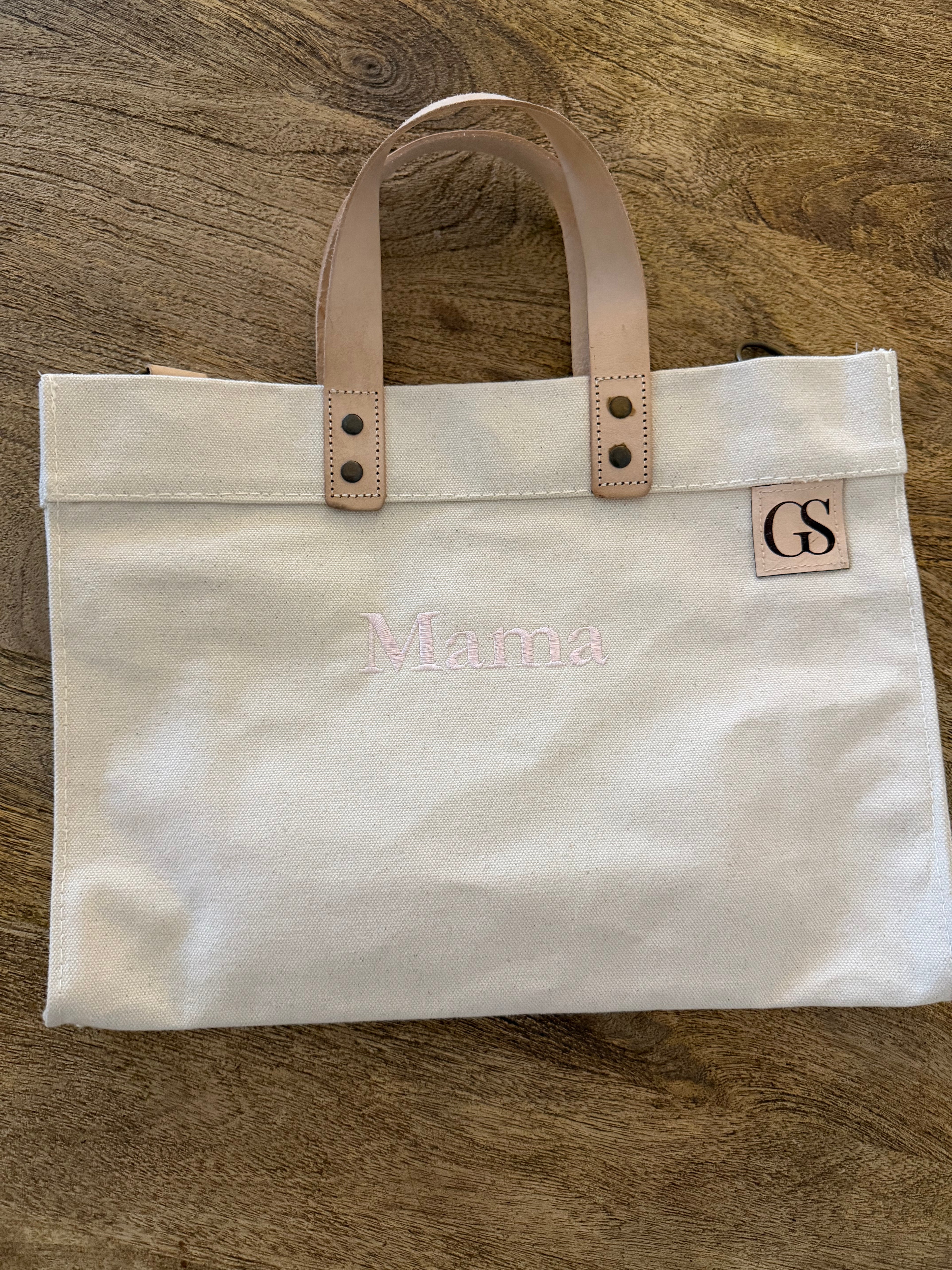 An off-white canvas tote bag with light tan leather handles and a small square leather patch with the initials 'GS'. The bag has 'Mama' embroidered in light pink across the front, and is photographed flat on a brown wood-grain surface.