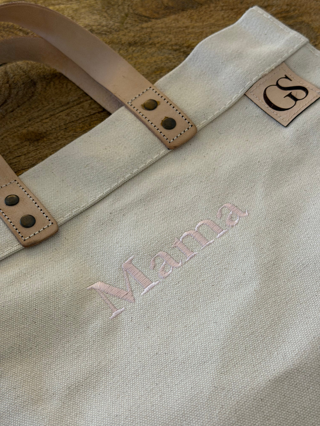 A close-up of a canvas tote bag with light tan leather handles and a small leather patch showing the initials 'GS'. The bag is personalized with the name 'Mama' embroidered in light pink thread, resting on a rustic wooden surface.