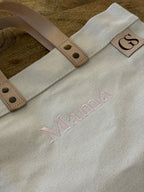 A close-up of a canvas tote bag with light tan leather handles and a small leather patch showing the initials 'GS'. The bag is personalized with the name 'Mama' embroidered in light pink thread, resting on a rustic wooden surface.