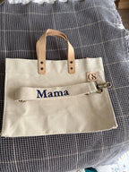 A beige canvas tote bag with tan leather handles and a matching canvas shoulder strap featuring the embroidered word 'Mama' in navy blue. A small leather patch on the bag is branded with 'GS'.