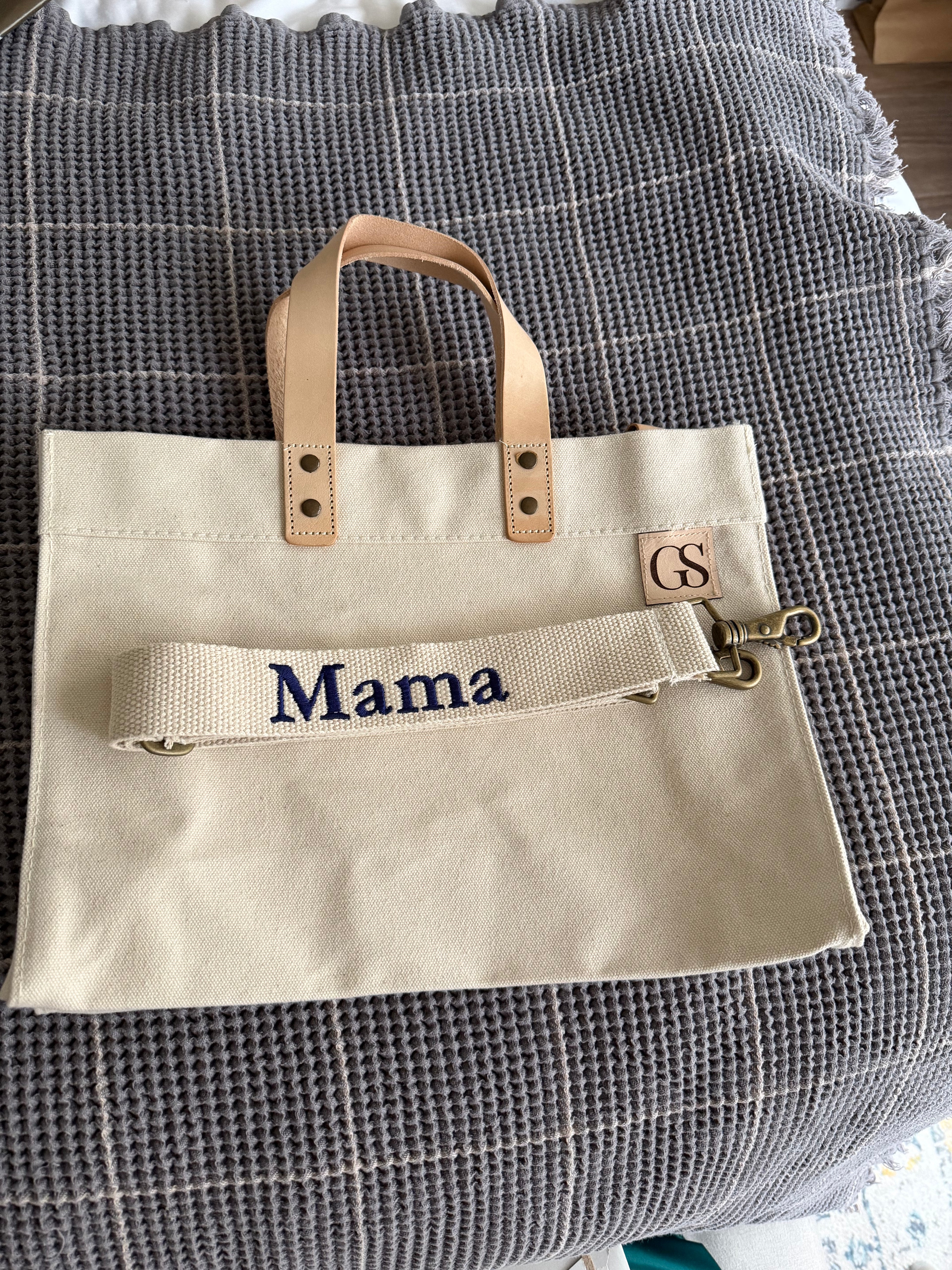 A beige canvas tote bag with tan leather handles and a matching canvas shoulder strap featuring the embroidered word 'Mama' in navy blue. A small leather patch on the bag is branded with 'GS'.
